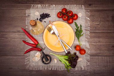 Taze domates, biber ve diğer baharatlar ve otlar ahşap masa ve bez peçete merkezinde modern sarı plaka çevresinde. Çatal ve bıçak plaka üzerinde bağlı. Üstten Görünüm. Yatay.