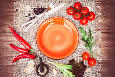 Fresh tomatoes, chili pepper and other spices and herbs around modern orange plate in the center of wooden table and cloth napkin. Top view. Blank place for your text. Horizontal.
