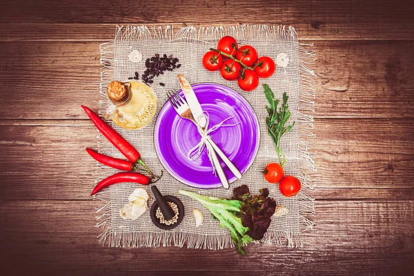 Taze domates, biber ve diğer baharatlar ve otlar ahşap masa ve bez peçete merkezinde modern mor plaka çevresinde. Çatal ve bıçak plaka üzerinde bağlı. Üstten Görünüm. Yatay.