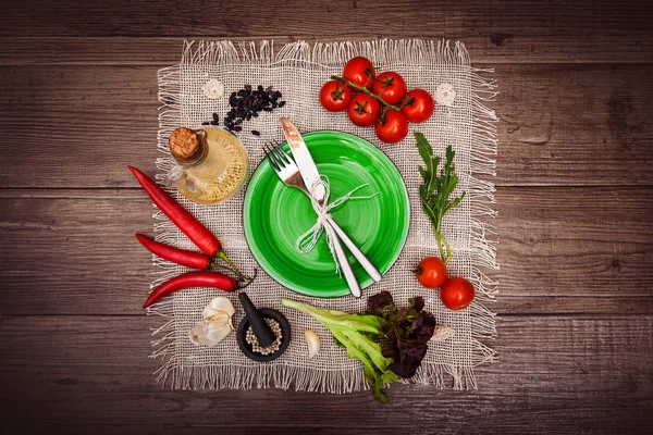 Taze domates, biber ve diğer baharatlar ve otlar ahşap masa ve bez peçete merkezinde modern yeşil plaka çevresinde. Çatal ve bıçak plaka üzerinde bağlı. Üstten Görünüm. Yatay.