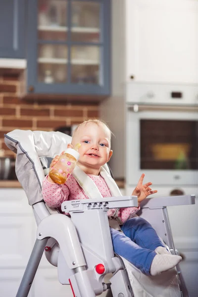 Einjähriges Kleinkind sitzt auf Kinderhochstuhl mit Fütterungsflasche in der Hand. — Stockfoto