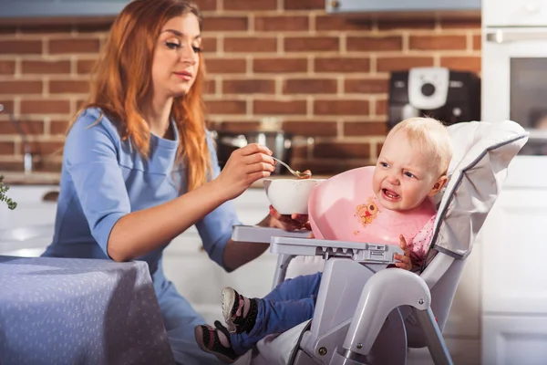 Little baby girl sits in high chair and feeding with spoon her beautiful mother