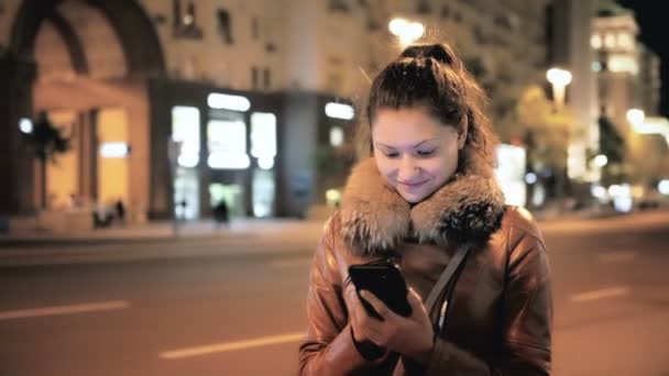 Chica feliz usando Smartphone contra el trasfondo de la ciudad nocturna — Vídeos de Stock