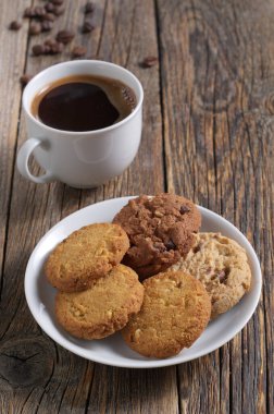 Beyaz tabakta çeşitli kurabiyeler ve tahta masada bir fincan kahve.