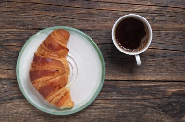 Kruvasan tabakta, kahve fincanı da ahşap arka planda.