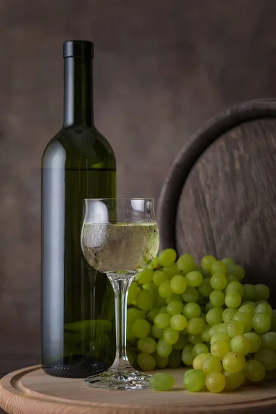 Una bottiglia di vino bianco, un bicchiere di vino bianco su fondo d'uva e botte sul tavolo di legno — Foto Stock