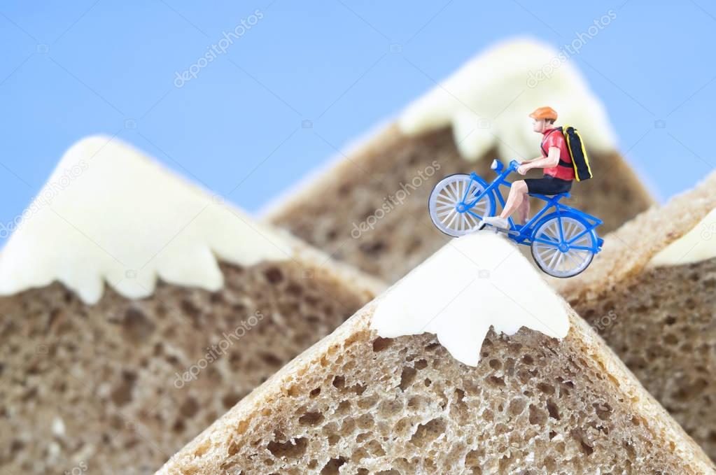 A toy cyclist is taking a ride on mountains, made of healthy rye bread and creamy cheese. Ecotourism and healthy eating concept.