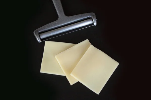 Slices of yellow cheese and a cheese slicer on black background. Top view close up image.