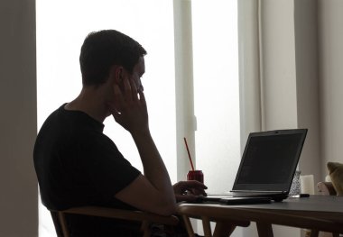 Evdeki genç adamın silueti elinde samanla dizüstü bilgisayarın yanında oturuyor..   