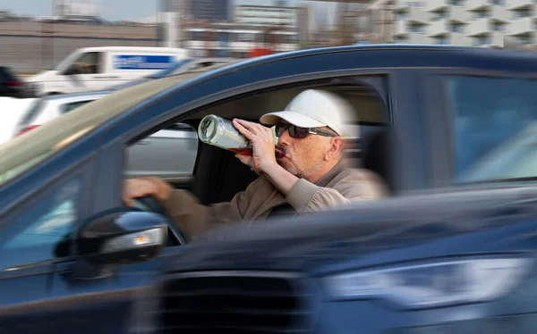 Careless male driver with sunglasses and cap drinking liquor from a bottle and driving in heavy traffic. Careless or reckless driving is a traffic violation. Motion blurred image with selective focus.