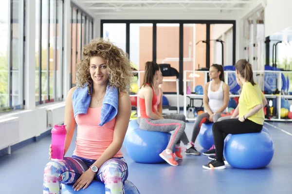 Hermosa joven sentada en la bola azul de pilates y sonriendo. Grupo de atletas femeninas sentadas detrás . — Foto de Stock