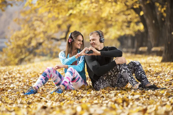 Bella giovane coppia che riposa e ascolta musica dopo un allenamento di successo nel parco. Cuore a forma di mano. Ambiente autunnale . — Foto Stock