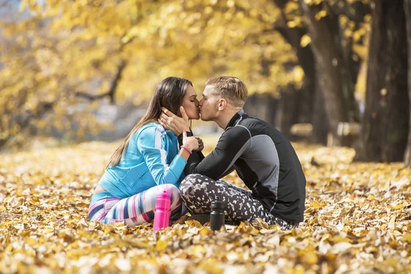 Schönes junges Paar, das sich nach dem erfolgreichen Training im Park ausruht und küsst. Herbstliches Umfeld. — Stockfoto