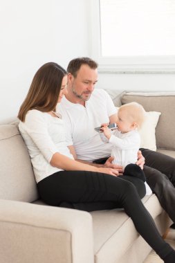 Üç mutlu aile evde keyif alıyor. Kanepede oturan ve cep telefonu ile oynarken onların güzel kız bebek bakarak anne. Aile değerleri. Boş zaman birlikte.