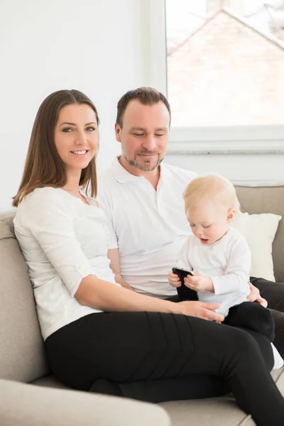 Famiglia felice di tre persone si sta divertendo a casa. I genitori seduti sul divano e guardando la loro bella bambina che gioca con il cellulare. Valori familiari. Tempo libero insieme . — Foto Stock