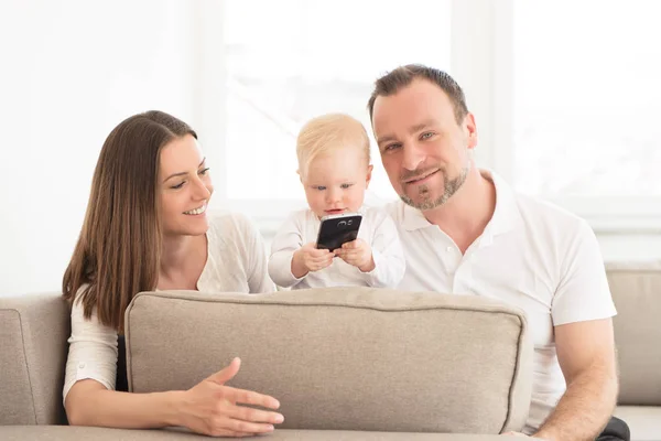 Famiglia felice di tre persone si sta divertendo a casa. Genitori seduti sul divano. La loro bellissima bambina che gioca con il cellulare. Valori familiari. Tempo libero insieme . — Foto Stock