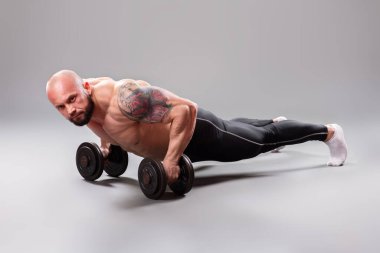 Seksi kaslı vücut bulucu gri arka planda ağırlık kaldırarak şınav çekiyor.