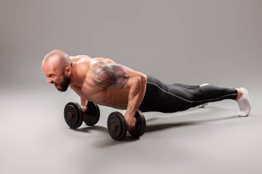 Seksi kaslı vücut bulucu gri arka planda ağırlık kaldırarak şınav çekiyor.