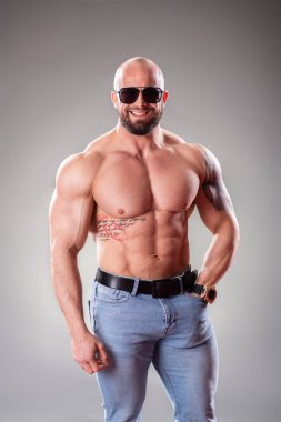 Gri arka planda duran güneş gözlüklü seksi kaslı vücut bulucu.