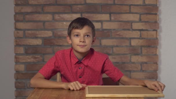 Niño sentado en el escritorio sosteniendo rotafolio con letras sí en la pared de ladrillo rojo de fondo . — Vídeo de stock