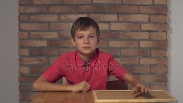 Kind sitzt am Schreibtisch und hält Flipchart mit Schriftzug Lächeln auf dem Hintergrund rote Backsteinwand. — Stockvideo