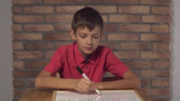 Enfant assis au bureau tenant tableau à feuilles avec signature de lettrage sur le mur de brique rouge arrière-plan . — Video