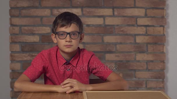 Niño sentado en el escritorio sosteniendo el rotafolio con letras disparadas en la pared de ladrillo rojo de fondo . — Vídeos de Stock