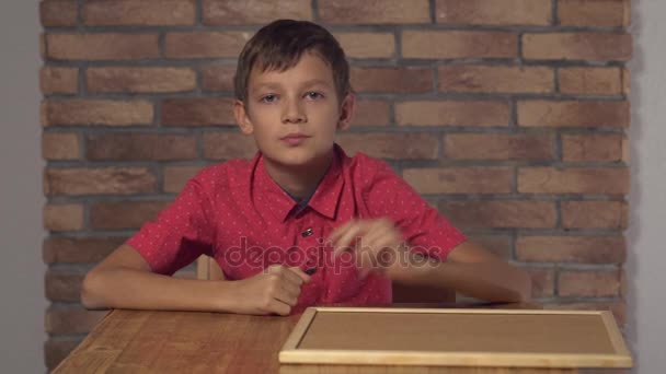 Niño sentado en el escritorio sosteniendo el rotafolio con letras me llaman en la pared de ladrillo rojo de fondo — Vídeos de Stock