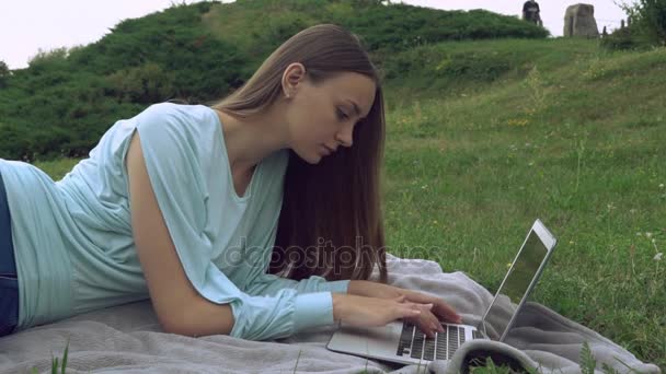 Μια όμορφη νεαρή γυναίκα είναι ξαπλωμένη ouside και εργάζεται σε έναν υπολογιστή. — Αρχείο Βίντεο