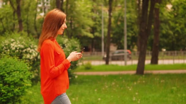 Lihat samping wanita berambut merah berjalan di jalan memegang smartphone — Stok Video