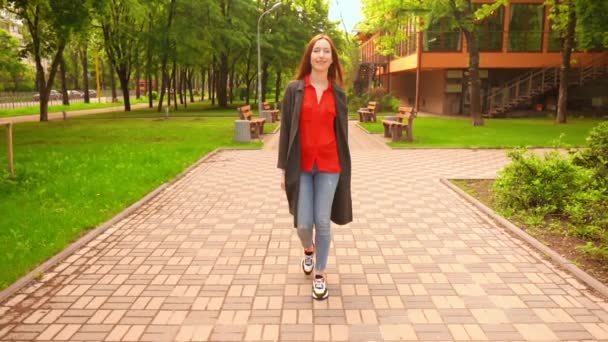 Рудоволоса жінка ходить сміючись у місті, оточеному зеленими деревами і травою — стокове відео