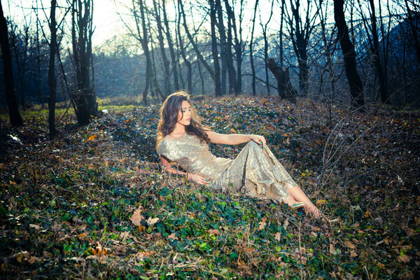 Beautiful young woman in long elegant dress sit on ground in forest