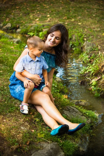 Молода усміхнена мати сидить на траві зі своїм маленьким сином — стокове фото