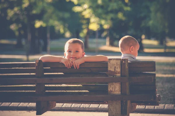 Dwóch małych chłopców braci siedzieć na drewnianej ławce w parku, letni dzień — Zdjęcie stockowe