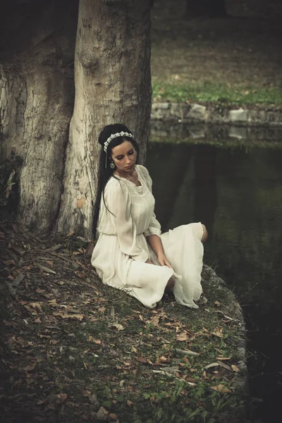 Молодая женщина в романтическом платье и украшения для волос сидеть у озера — стоковое фото