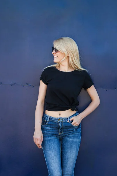 身着休闲装的黑色T恤 戴着太阳镜 头戴蓝色金属背景的金发女青年在城市T恤造型 — 图库照片