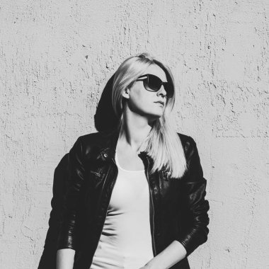 Deri ceketli ve güneş gözlüklü genç sarışın kadının siyah beyaz portresi şehir yaz gününde duvarda asılı duruyor.