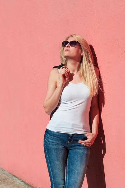 年轻的金发女郎 戴着太阳镜 头戴白色的背心 靠着粉红的墙壁 炎热的夏天 在城市里晒太阳 — 图库照片