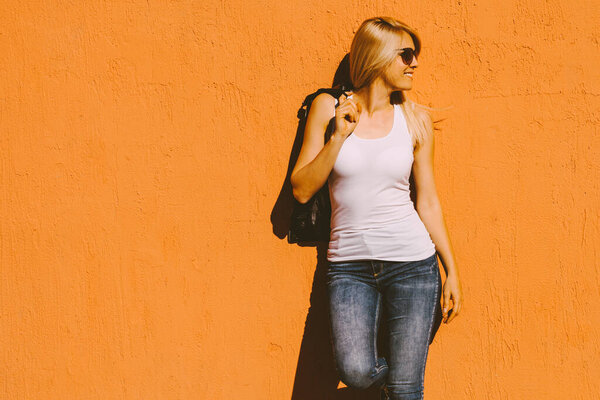 young  smiling blonde woman in white tank top and sunglasses against yellow wall outdoor in city summer heat 