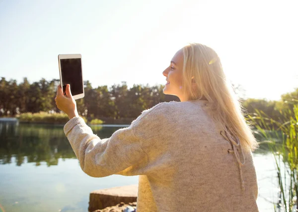 Dívka v podzimním parku nedaleko jezera drží tablet — Stock fotografie