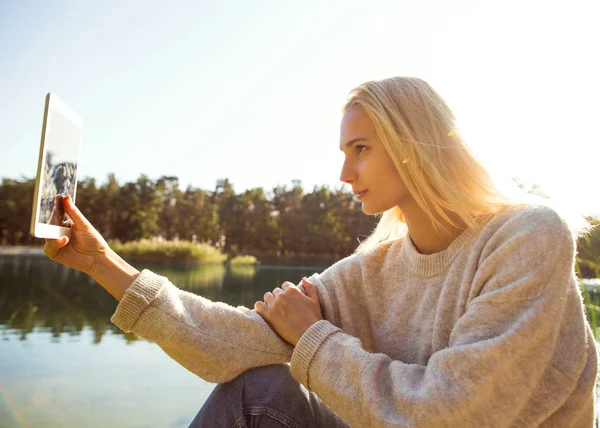 Meisje in een herfst park in de buurt van een meer houdt een tablet — Stockfoto