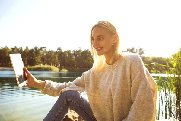 Dívka v podzimním parku nedaleko jezera drží tablet — Stock fotografie