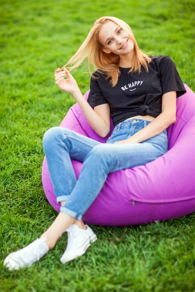 Flickan sitter på gräset i parken på stol väska — Stockfoto