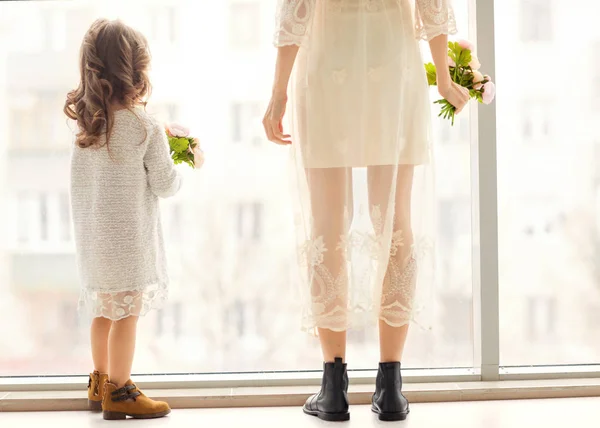 Madre e hija se divierten de pie cerca de una enorme ventana, la hija le da flores a su madre en el día de su madre . —  Fotos de Stock