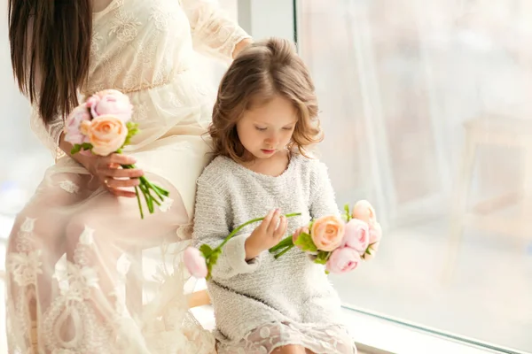 Madre e hija se divierten de pie cerca de una enorme ventana, la hija le da flores a su madre en el día de su madre . — Foto de Stock