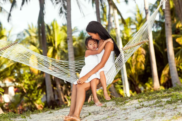 asian happy mother with her daughter sitting in a hammock on the shore of a tropical beach. Mothers day.