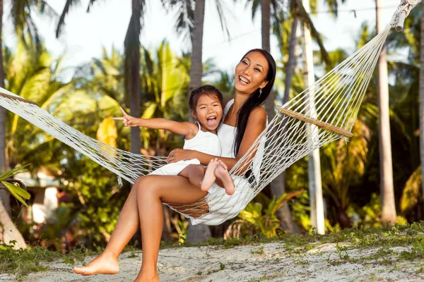 Ασιατικές ευτυχισμένη μητέρα με την κόρη της κάθεται σε μια αιώρα στην ακτή της μια τροπική παραλία. Ημέρα της μητέρας. — Φωτογραφία Αρχείου