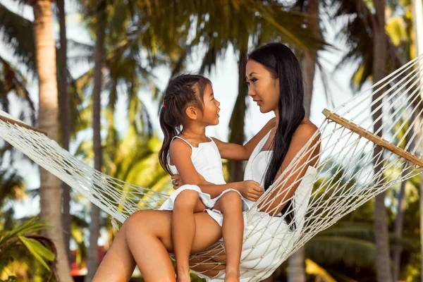 asian happy mother with her daughter sitting in a hammock on the shore of a tropical beach. Mothers day.