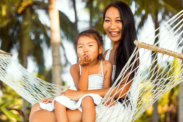 Mãe feliz asiática com sua filha sentada em uma rede na costa de uma praia tropical. Dia das mães . — Fotografia de Stock