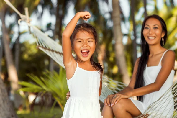 asian happy mother with her daughter sitting in a hammock on the shore of a tropical beach. Mothers day.